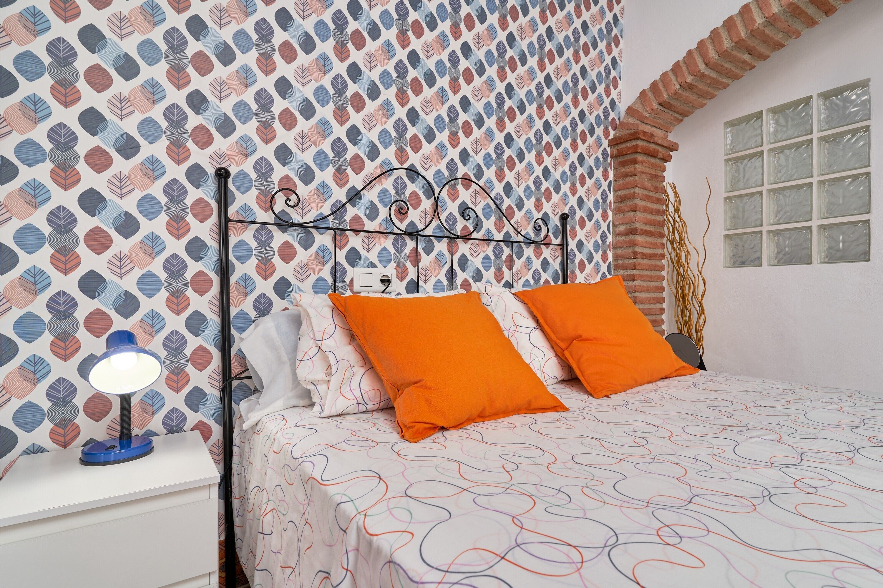 Une chambre avec un papier peint à motifs, un cadre de lit en métal, deux coussins orange, une table de chevet blanche avec une lampe, et une fenêtre surmontée d’une arche en briques.
