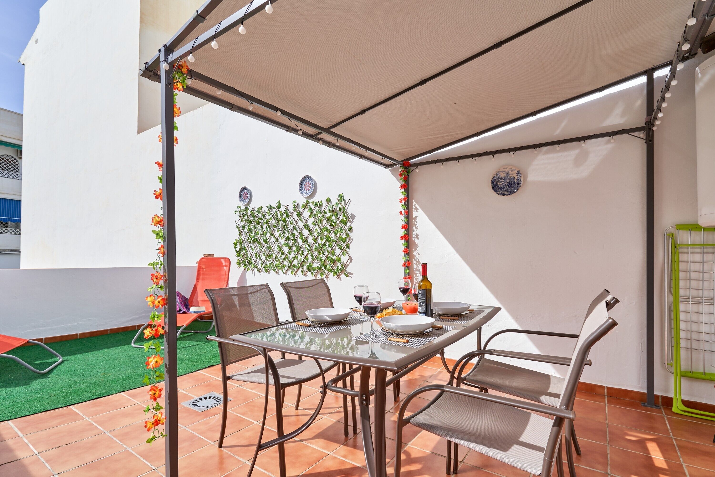 Una terrazza con un tavolo apparecchiato per mangiare, circondata da sedie, in una zona ombreggiata con una pianta decorativa.