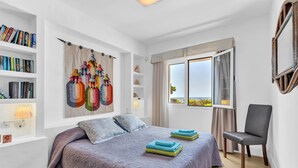 Un dormitorio con una cama, una silla, una estantería, un ventanal con vistas y una pared decorada con una obra de arte colorida.