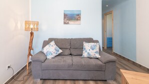Un canapé gris agrémenté de deux coussins à motifs, un tableau accroché au mur, un lampadaire en bois et un couloir avec un mur bleu et une porte en bois.