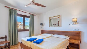 Una cama doble con dos toallas azules, un cabecero de madera, una mesita de noche, un ventilador de techo, una ventana con cortinas, un espejo montado en la pared y una lámpara de pared.