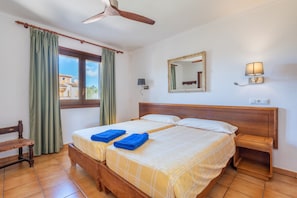 A double bed with two blue towels, a wooden headboard, a nightstand, a ceiling fan, a window with curtains, a wall-mounted mirror, and a wall lamp. - Apartment 'Pedrassa Gran Superior' with Sea View, Shared Pool and Wi-Fi (Se Torre Nova)