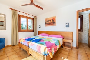 Un dormitorio con una cama de madera, un ventilador de techo, una ventana con cortinas, un cuadro en la pared y una mesita de noche de madera.