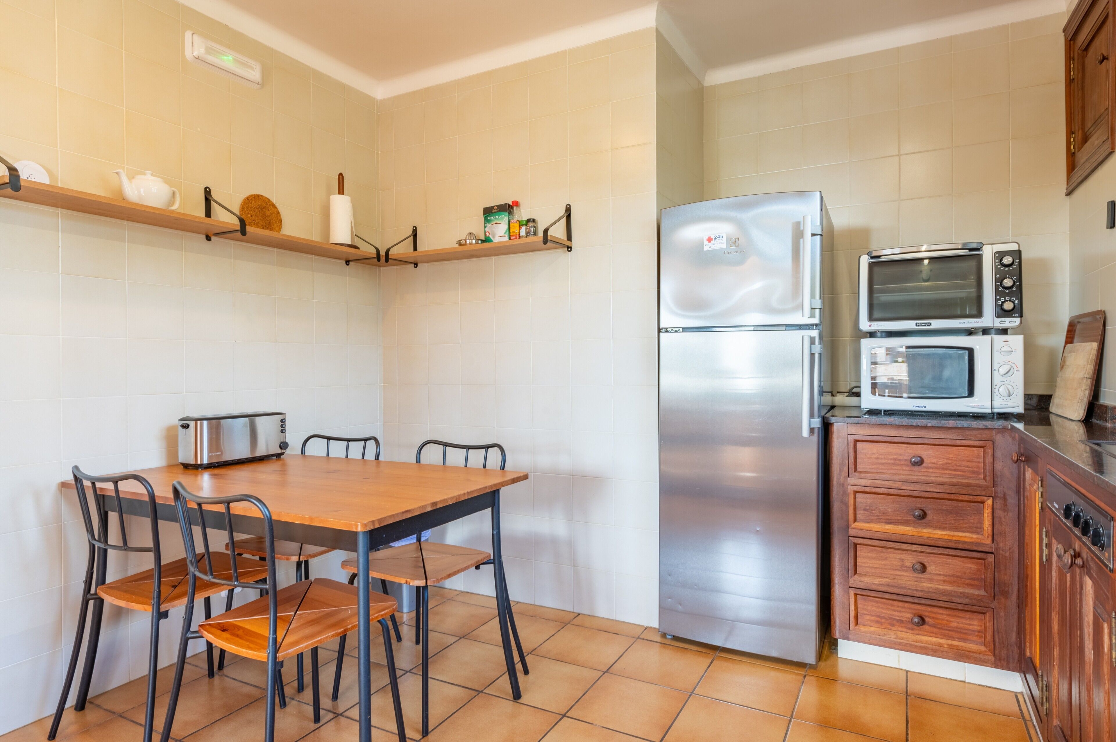 Una cocina con una mesa de madera, sillas, un refrigerador, un microondas y una estantería con utensilios de cocina.