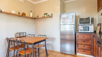 Una cocina con una mesa de madera, sillas, un refrigerador, un microondas y una estantería con utensilios de cocina.