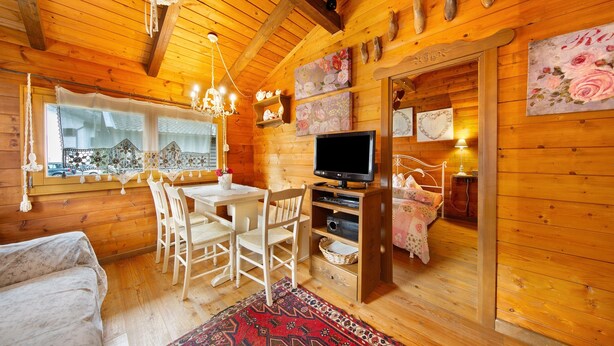 L'intérieur d'un chalet en bois comprend une table à manger, des chaises, un téléviseur, un lit et un tapis rouge à motifs.