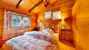 Une chambre à coucher dans un chalet en bois, avec un lit, des rideaux en dentelle aux fenêtres, des lampes fixées au mur et des œuvres d’art encadrées.