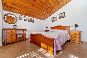 A bedroom with a wooden ceiling, a bed with a patterned bedspread, a wooden dresser, a nightstand with a lamp, and a wall-mounted mirror.