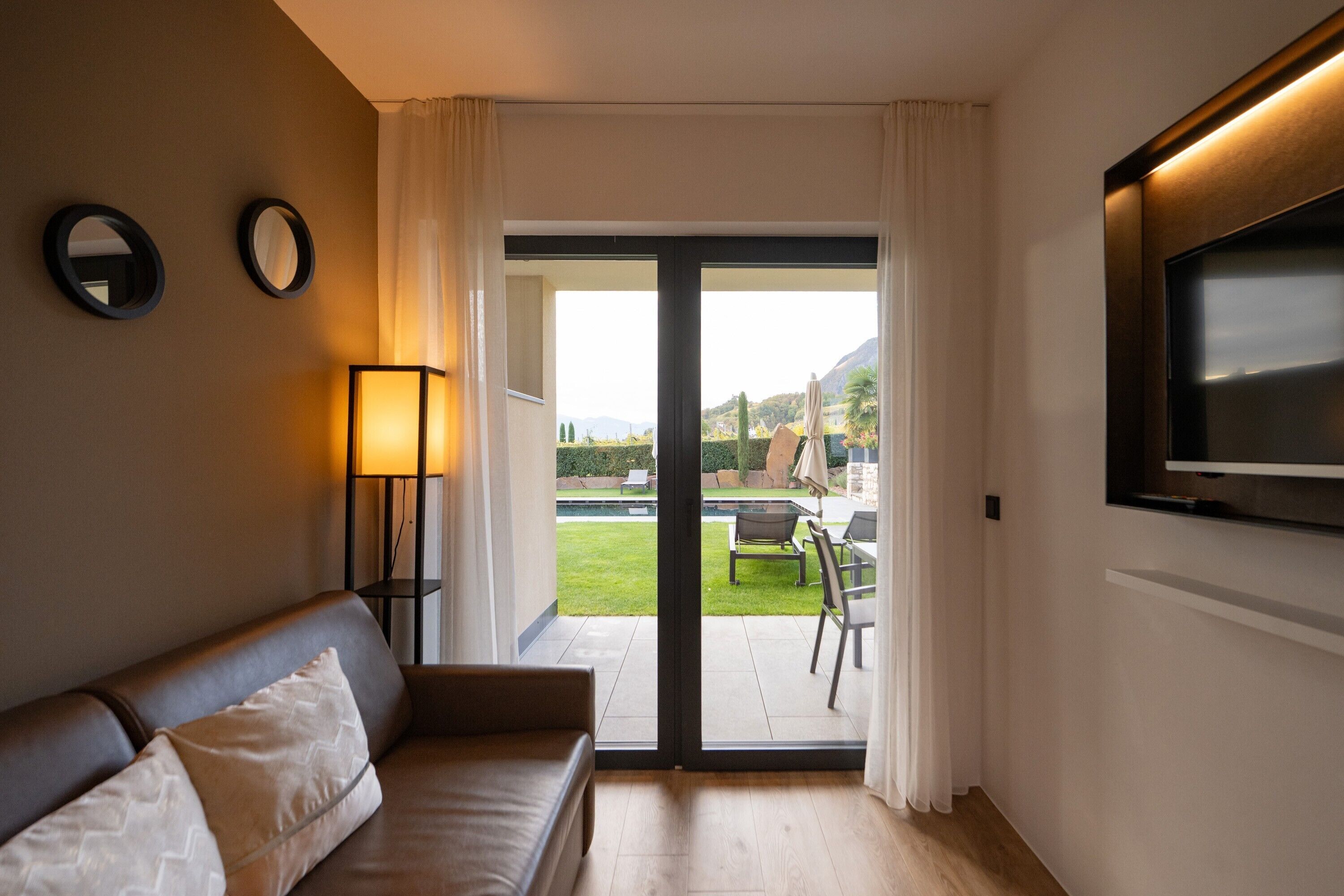 Een kamer met een bank, een aan de muur bevestigde televisie, en een schuifpui naar een buitenruimte met een zwembad en tuinmeubilair.