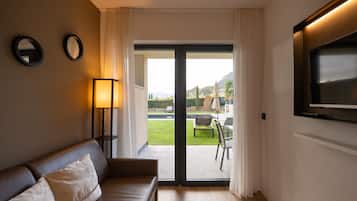 Een kamer met een bank, een aan de muur bevestigde televisie, en een schuifpui naar een buitenruimte met een zwembad en tuinmeubilair.