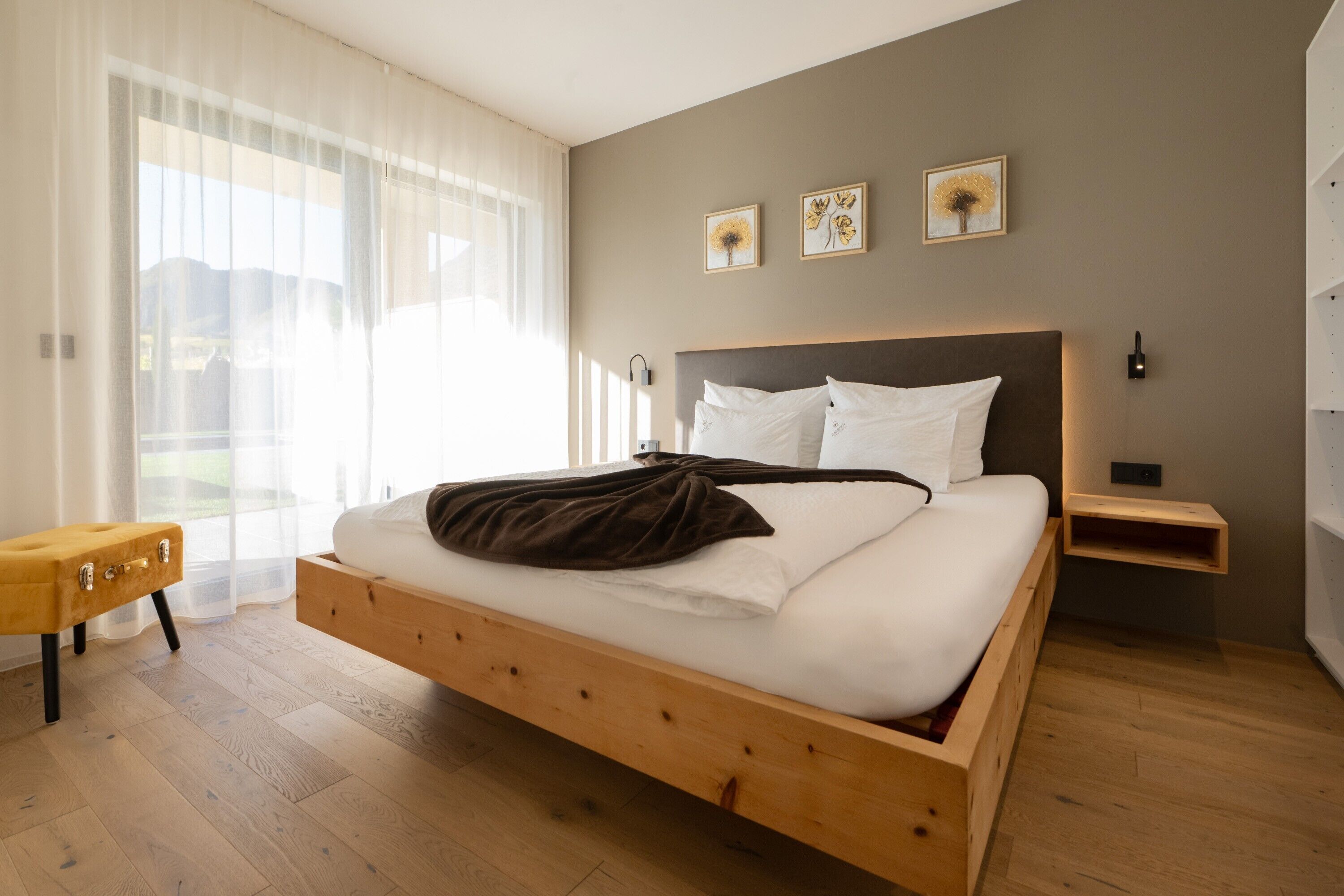Une chambre à coucher moderne avec un lit en bois, une table de chevet, un petit banc en bois et une grande fenêtre ornée de rideaux légers.