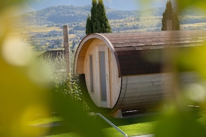 Un bâtiment en forme de tonneau, fait de bois, avec une porte et des fenêtres, entouré d’une zone herbeuse et d’arbres, et offrant une vue sur des collines en arrière-plan.