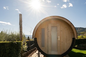 Un edificio con forma de barril de madera, con puerta y ventanas, rodeado de vegetación y montañas.