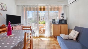 Ein Zimmer mit Fernseher, einem Tisch mit einem Tischläufer mit Sternenmuster, einer Couch mit gemusterten Kissen, einer Kaffeemaschine und einer Klimaanlage.