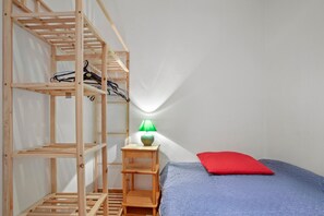Une chambre avec un lit superposé, une étagère en bois, une petite table avec une lampe et un oreiller rouge.