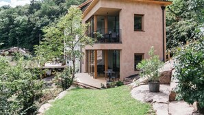 Ein modernes, zweistöckiges Haus mit Balkon, umgeben von viel Grün und einer Bergkulisse.
