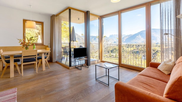 Een moderne woonkamer met een houten vloer, een bank, een televisie, een eettafel met stoelen en grote ramen met uitzicht op bergen.