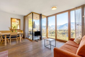 A modern living room with a wooden floor, a sofa, a TV, a dining table with chairs, and large windows offering a mountain view. - Apartment 'Firmelein 1 Stock' with Mountain View, Private Terrace and Wi-Fi (Missian)
