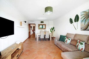 A living room with a sofa, a TV, a dining table with chairs, and a kitchen area in the background.
