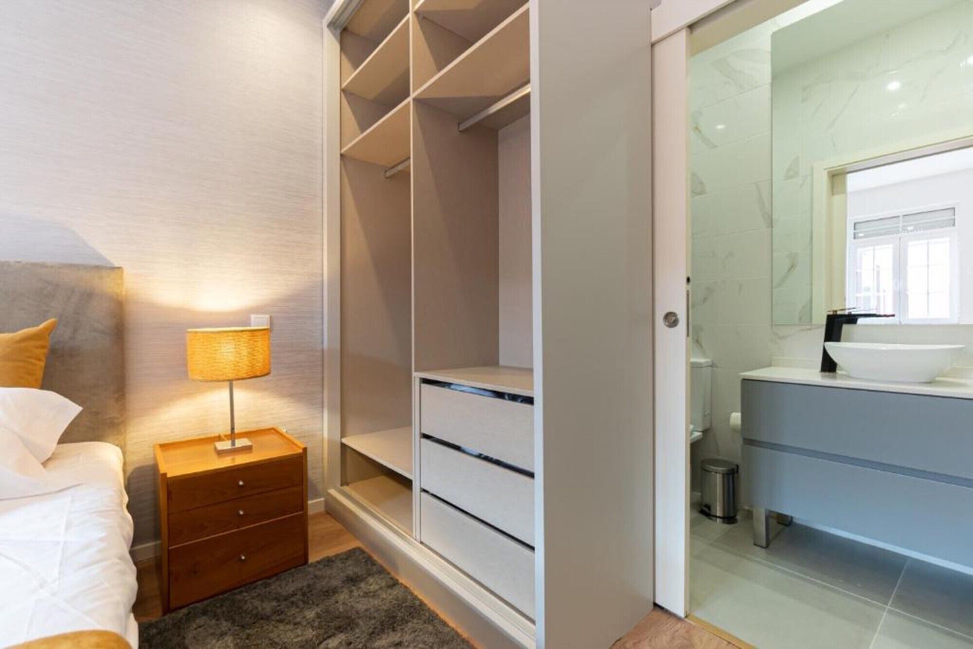 Ein Schlafzimmer mit Bett, Nachttisch, Lampe und einem begehbaren Kleiderschrank mit Schubladen.