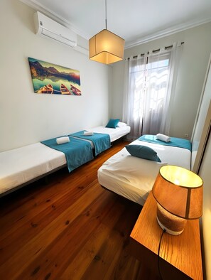 A room with two single beds, a wooden floor, a painting on the wall, and a hanging light fixture.