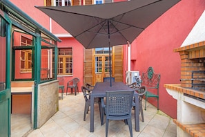 Um pátio externo com mesa e cadeiras, churrasqueira e um edifício vermelho ao fundo.