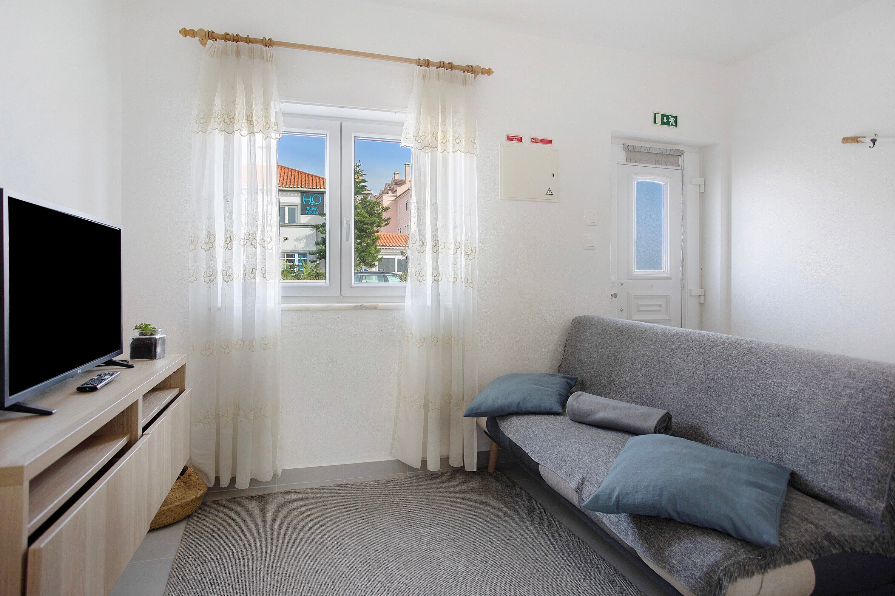 A room with a sofa, a television on a wooden stand, a window with curtains, and a door with an emergency exit sign.