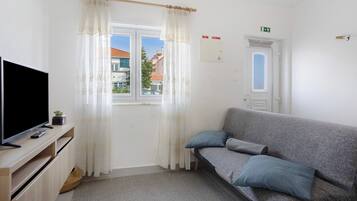 A room with a sofa, a television on a wooden stand, a window with curtains, and a door with an emergency exit sign.