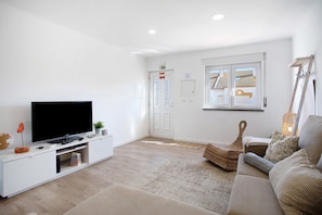 Una sala de estar con televisor de pantalla plana, paredes blancas, suelo de madera, sofá, mesa de centro y ventana con vista a edificios.