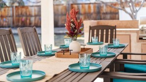 Un espace de restauration en plein air avec une table en bois dressée pour six personnes, agrémentée de verres et d’assiettes de couleur turquoise, d’un vase blanc contenant des fleurs rouges et d’un set de table en fibres tressées.