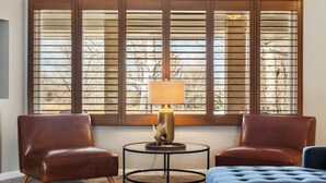 Une pièce comprenant deux fauteuils en cuir, un pouf bleu, une petite table ronde avec une lampe et une grande fenêtre avec des stores.