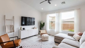 A modern living room with a ceiling fan, a flat-screen TV mounted on the wall, a white radiator, a large window with a view, and a patterned rug.