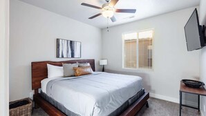 Une chambre avec un lit, un ventilateur de plafond, une fenêtre avec des stores, un téléviseur fixé au mur et une petite table.