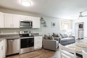 A modern kitchen with white cabinets, a microwave, stove, and dishwasher. There is a grey sofa and a dining table with chairs.