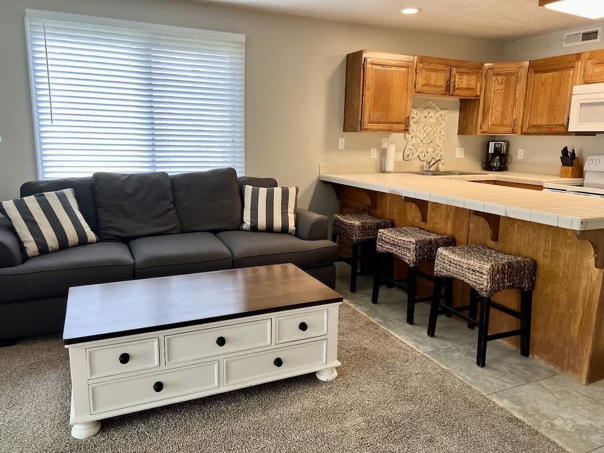 A living room with a dark sofa, a white coffee table with drawers, and a kitchen area with wooden cabinets and a bar stool.