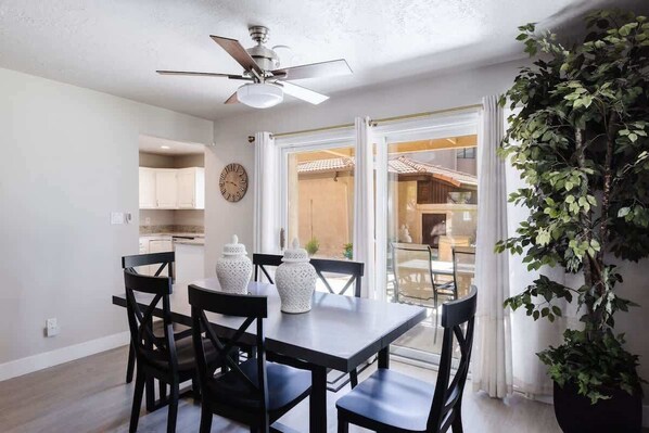 A dining area with a table and chairs, a ceiling fan, a clock on the wall, and a view through a glass door to the outside.