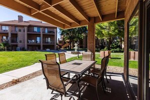 A covered outdoor patio with a table and chairs, a view of a building with balconies, and a grassy area with trees.
