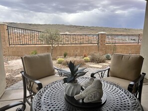 An outdoor patio with two chairs and a table, a cactus, and a view of a desert landscape.