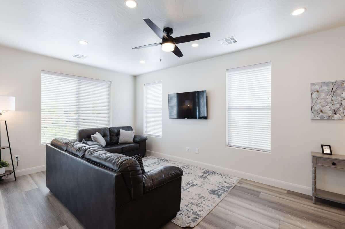 Un salon moderne avec un canapé en cuir foncé, un ventilateur de plafond et une grande fenêtre équipée de stores.