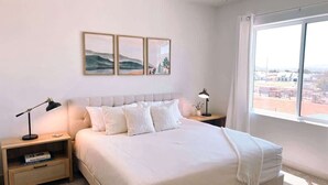 A bedroom with a large bed, two bedside tables with lamps, a window with a view, and three framed pictures on the wall. - Lava Flow @ Heritage Point - Santa Clara (Santa Clara)