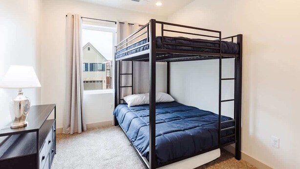 Une chambre avec un lit superposé, un ventilateur de plafond, un bureau avec une lampe et une vue sur un immeuble par la fenêtre.