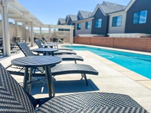 Zona de piscina con sillones y mesas, una piscina y casas modernas al fondo.