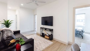 A modern living room with a flat-screen TV mounted on the wall, a black console table, a leather sofa, a red vase with a plant, and a ceiling fan. - Lava Flow @ Heritage Point - Santa Clara (Santa Clara)