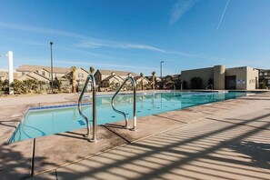 A swimming pool with metal ladders, surrounded by a concrete deck and palm trees, with residential buildings in the background.