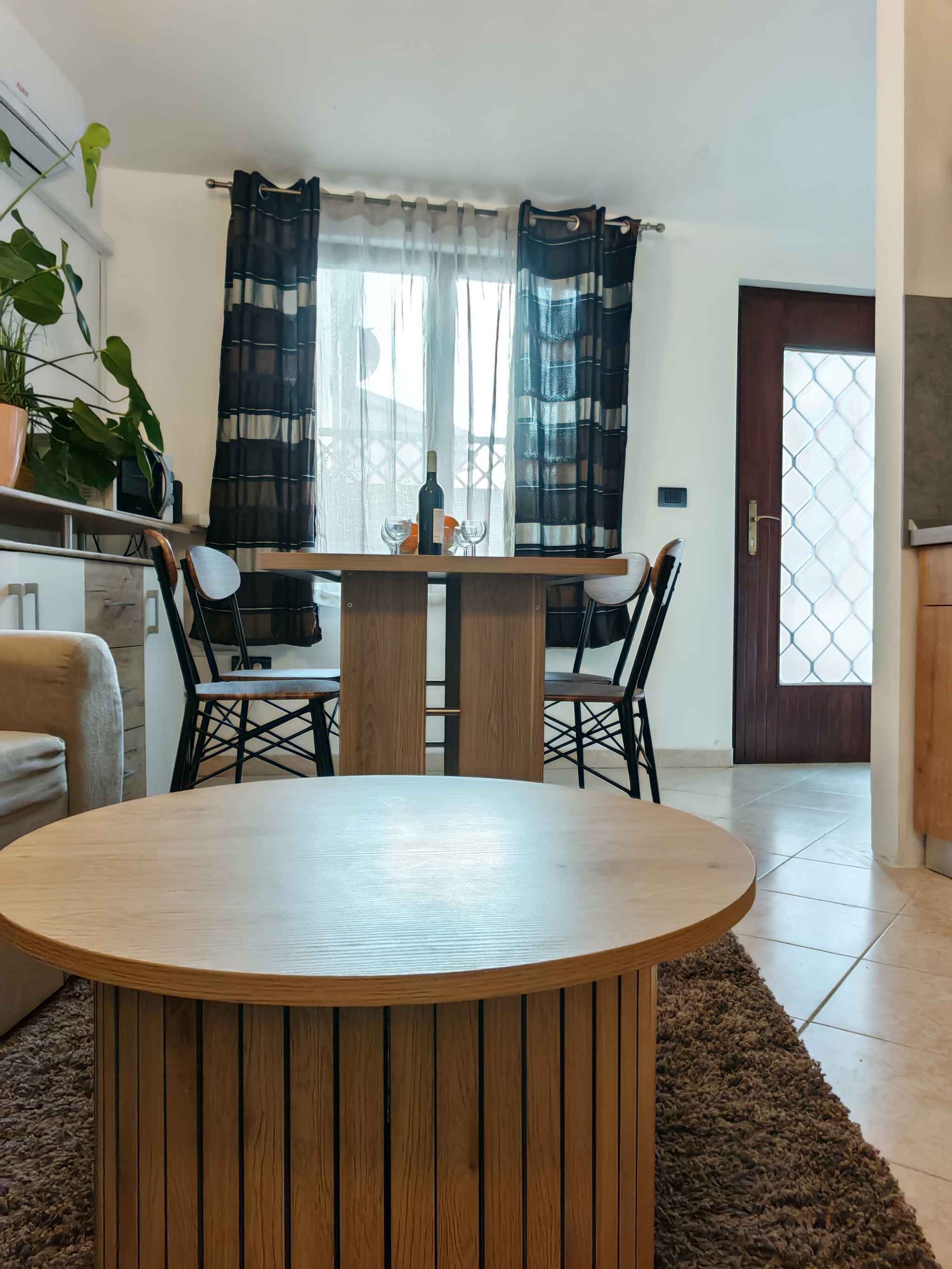 A dining area with a round wooden table, two chairs, a potted plant, and a window with curtains.