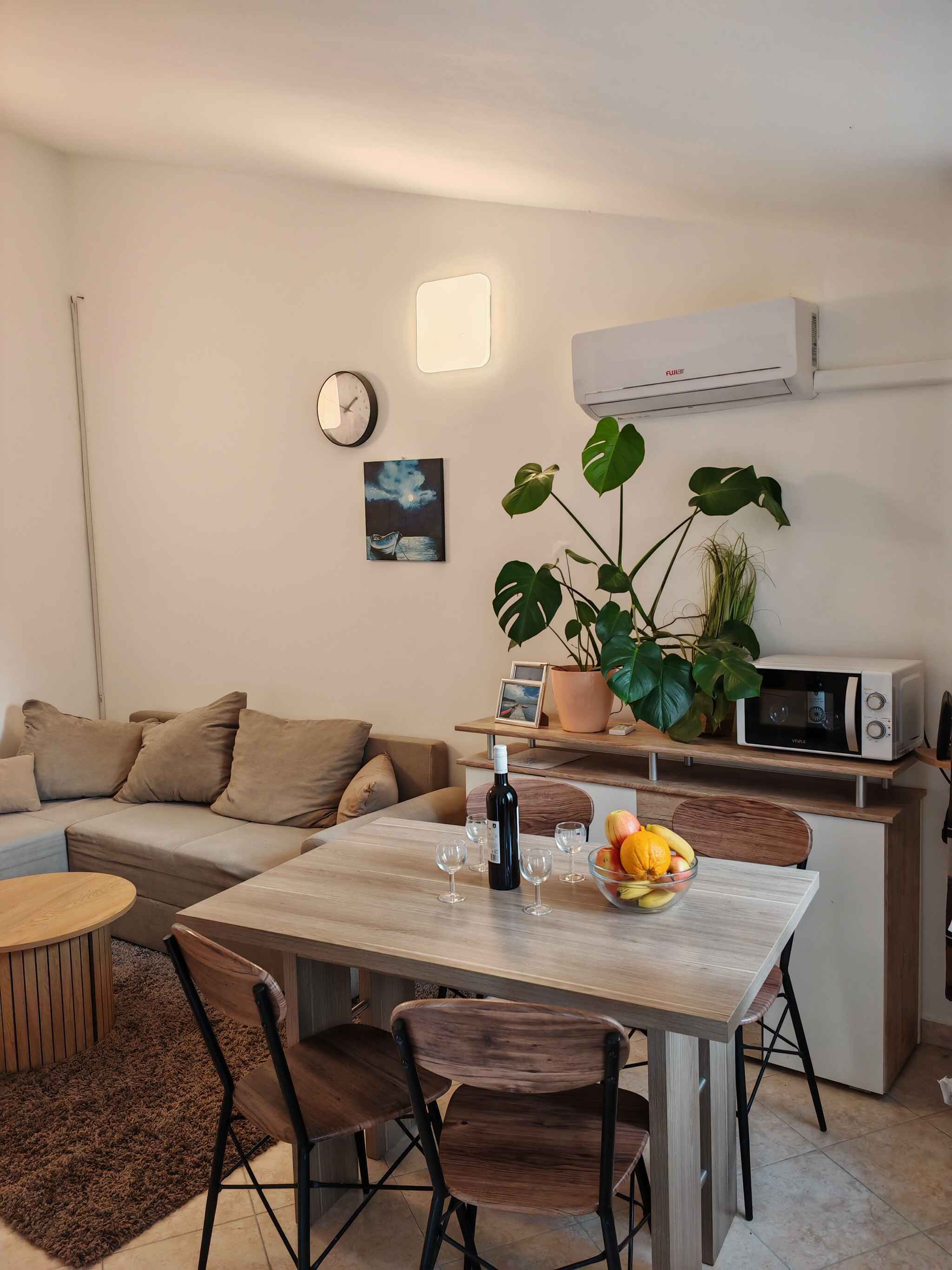 Un salon moderne comprenant un canapé, une table à manger, des chaises, une horloge, un tableau, une plante en pot, un four à micro-ondes et une bouteille de vin.
