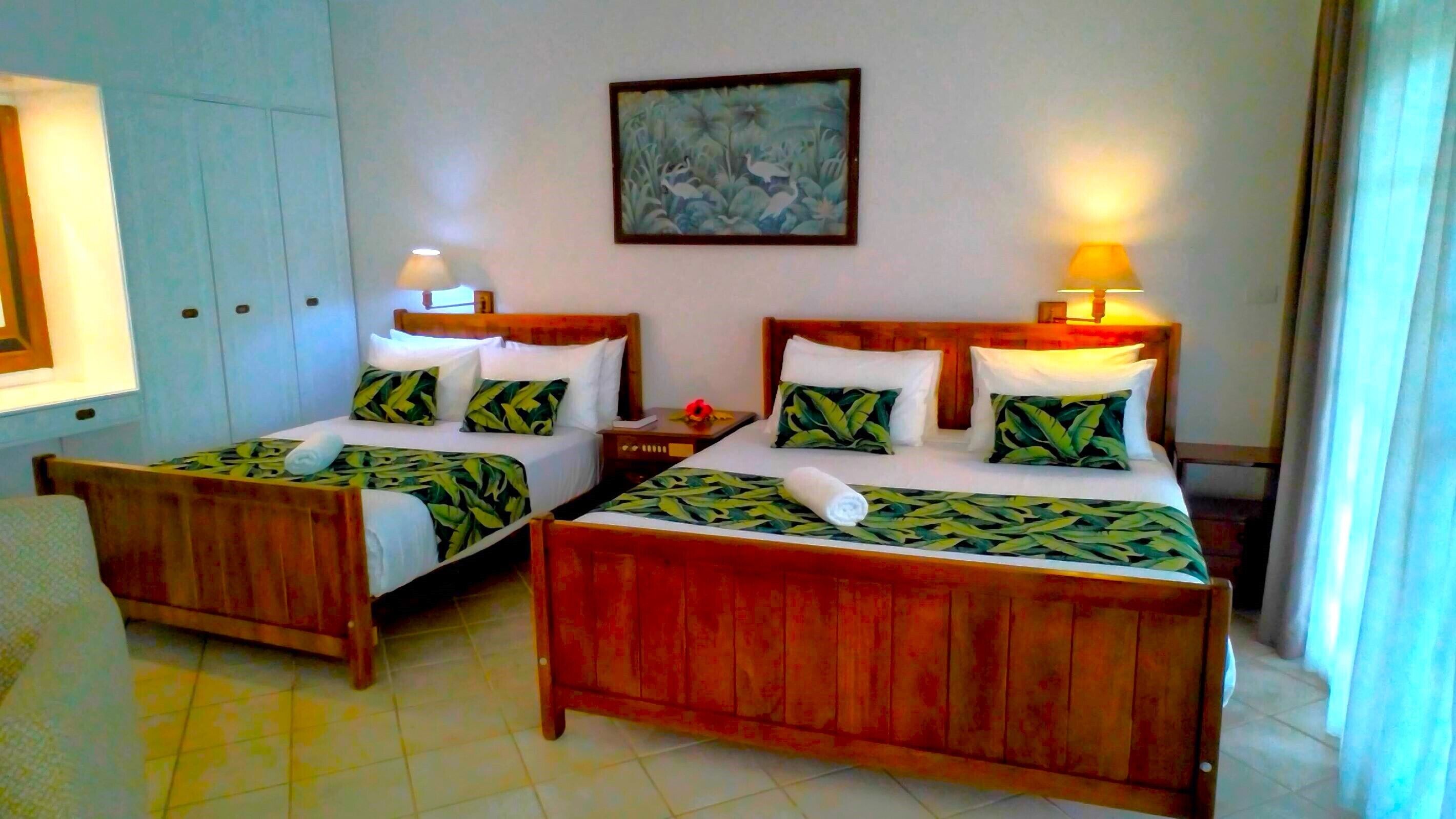 Una habitación con dos camas de madera, cada una con un edredón y almohadas a juego con estampado de hojas verdes. Hay un cuadro en la pared sobre las camas y una lámpara en cada mesita de noche.
