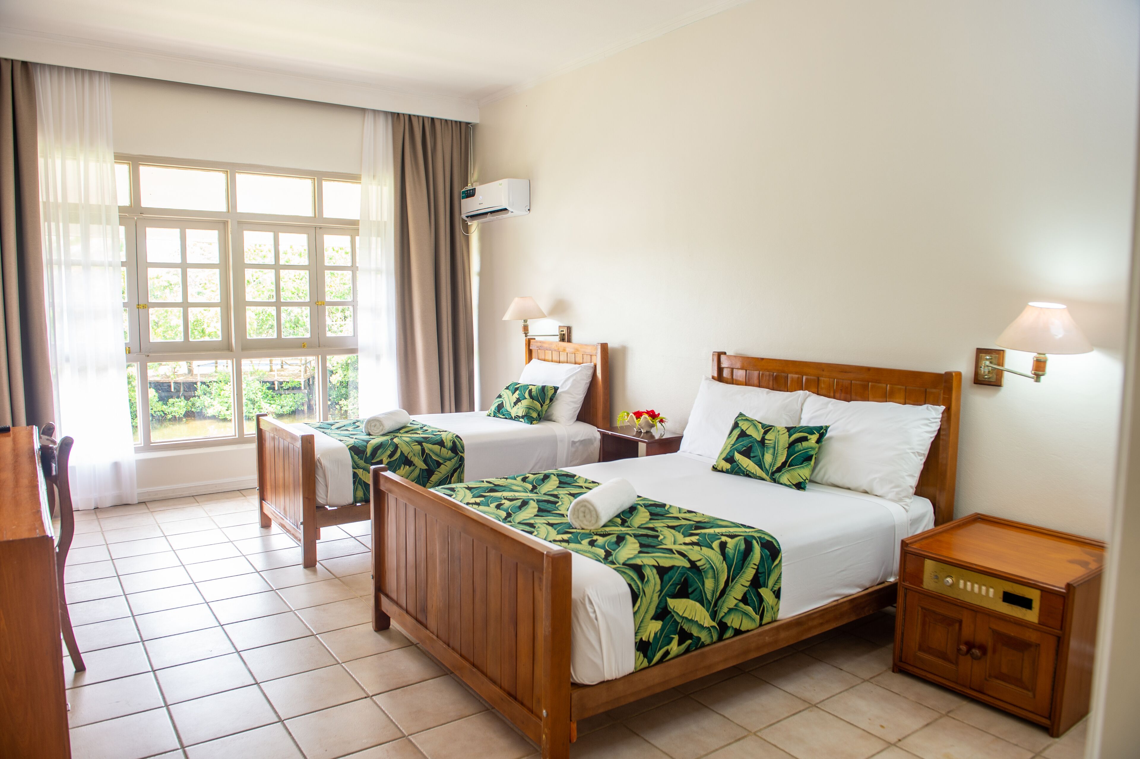 Une chambre d’hôtel avec deux lits en bois, une grande fenêtre avec des rideaux, une table de chevet avec une lampe et une petite armoire en bois.