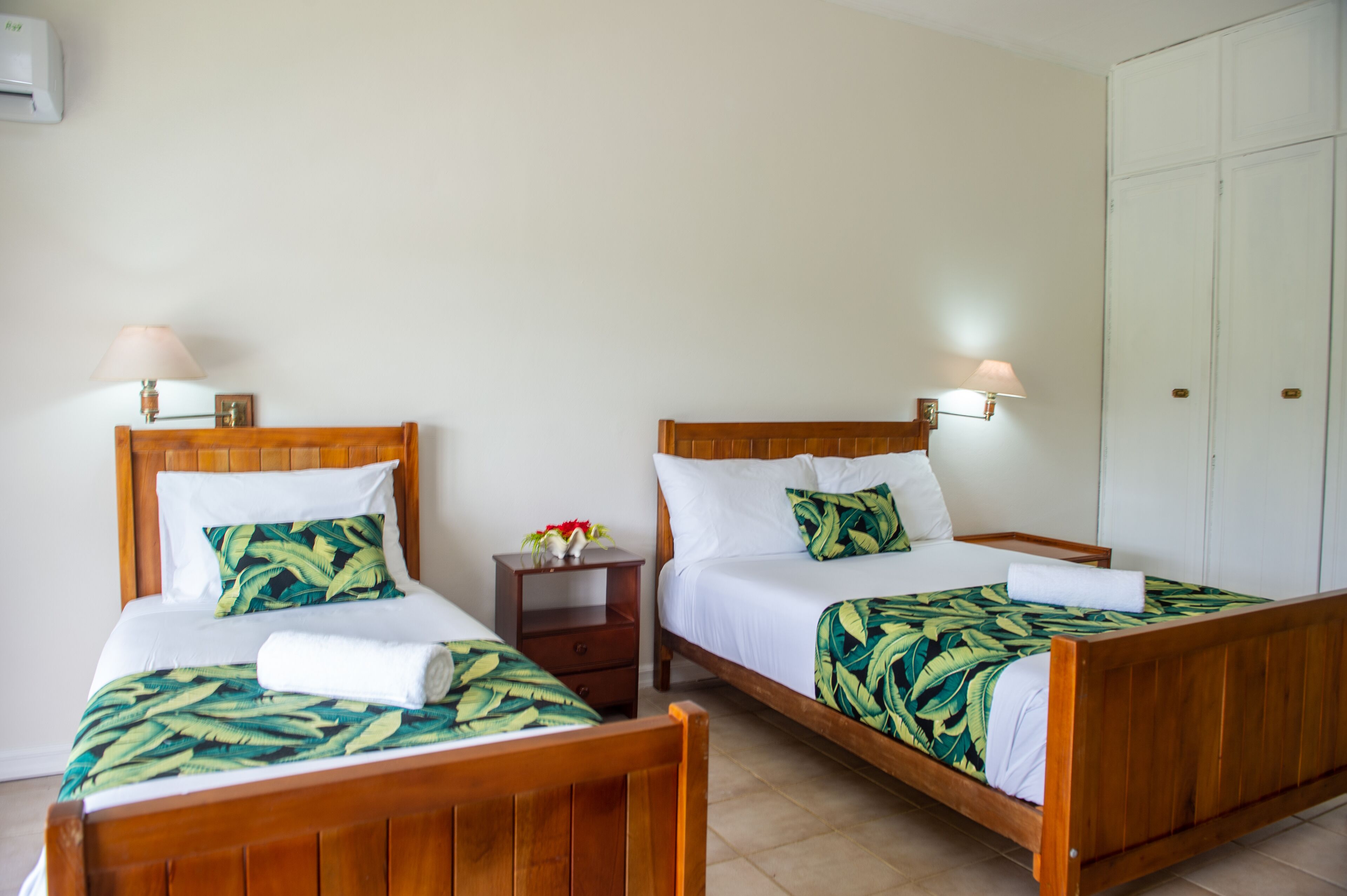 Two wooden beds with white linens and tropical leaf-patterned blankets, a small bedside table with a lamp, and a white wardrobe.
