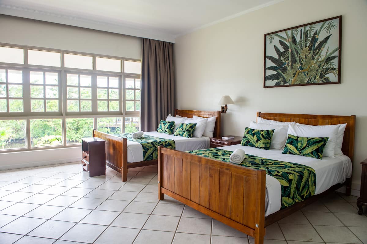 Una habitación de hotel con dos camas de madera, un ventanal con cortinas, un cuadro en la pared y suelo de baldosas.